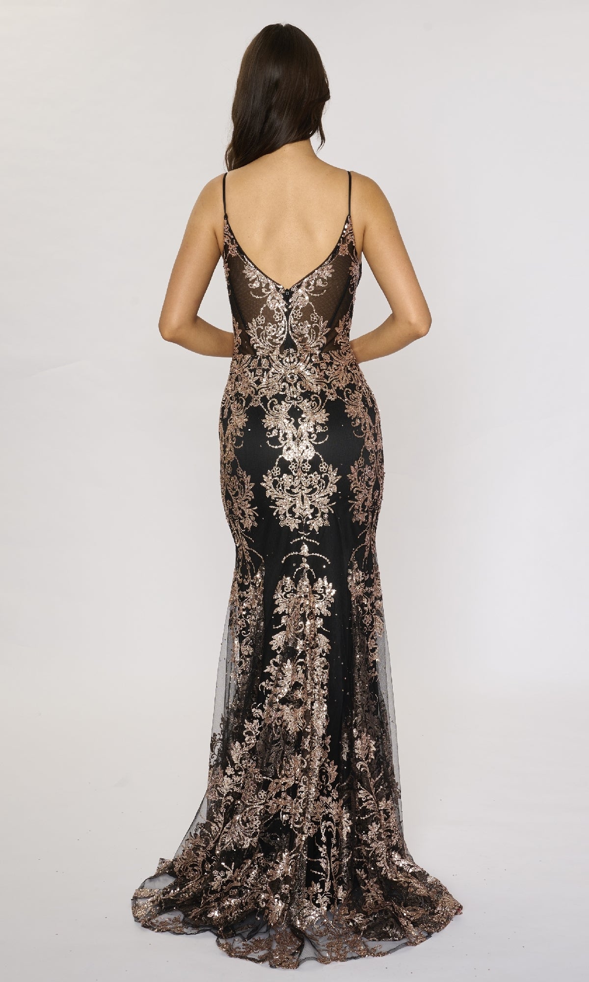 A woman faces away, wearing the Nina Canacci 1562 long formal dress—a black mermaid gown with gold floral embroidery, thin straps, and a sheer back detail.